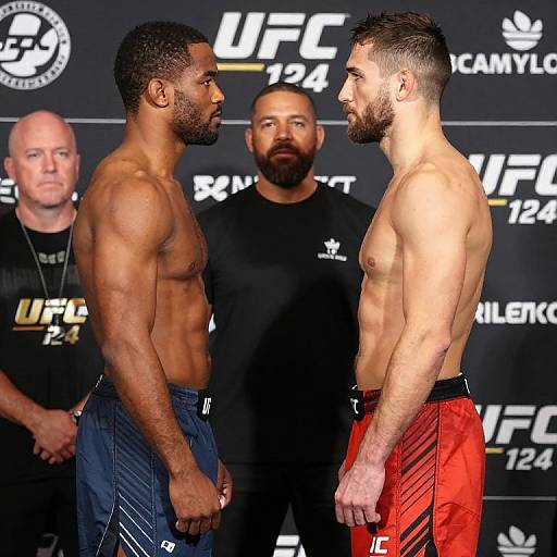 Photograph of two shirtless, muscular male UFC fighters facing each other, wearing blue and red shorts, with a bearded man and bald man in