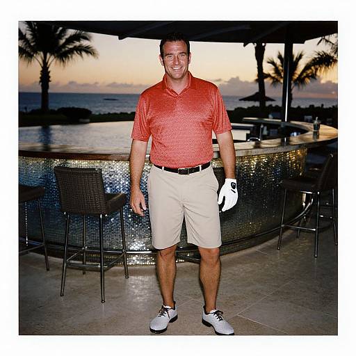 Photograph of a smiling man in a red polo, beige shorts, white sneakers, and golf glove, standing at a tropical bar with sunset and palm
