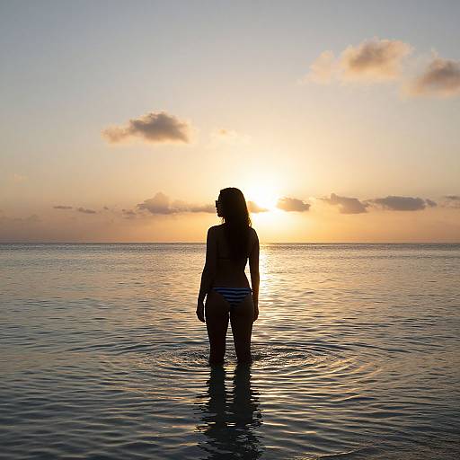Silhouetted woman in striped bikini stands in calm ocean at sunset, ripples around her, golden sky with scattered clouds.