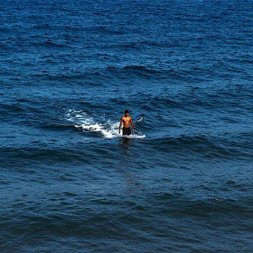Photograph of a lone male surfer wearing an orange shirt, standing on a small wave in deep blue ocean water.