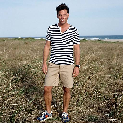 Photograph of a smiling man with short black hair, wearing a black-and-white striped shirt, beige shorts, and blue sneakers, standing in a grass
