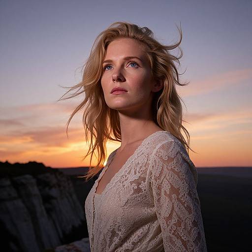 Photograph of a blonde woman with blue eyes, wearing a white lace top, standing against a sunset-lit cliff backdrop.