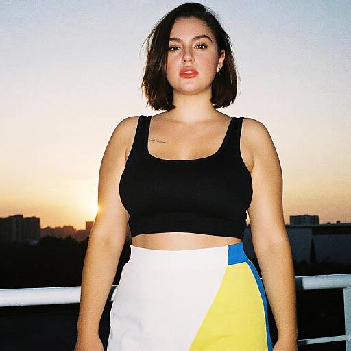 Photograph of a young woman with short brown hair, wearing a black sports bra and white, blue, and yellow high-waisted skirt, standing