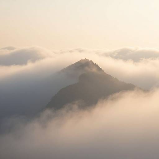 Photograph of a mist-covered mountain peak at sunrise, with soft, golden light illuminating the clouds and partially revealing the dark, shadowed slopes.