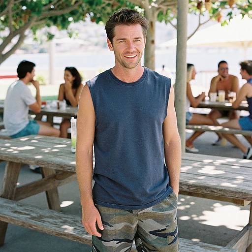 Photograph of a smiling, muscular man with short brown hair, wearing a sleeveless navy shirt and camo pants, standing outdoors at a sunlit