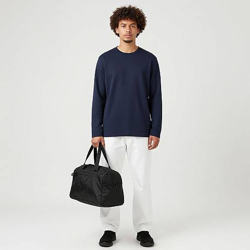Photograph of a curly-haired man in a navy sweatshirt, white pants, black shoes, holding a black duffel bag, against a white
