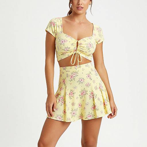 Photograph of a young woman with light skin wearing a yellow floral crop top and matching skirt, standing against a plain white background.
