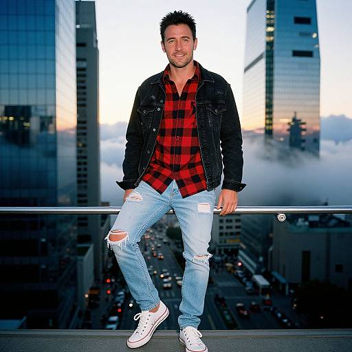 Photograph of a smiling, dark-haired man in a red plaid shirt, black jacket, ripped jeans, and white sneakers, standing on a city