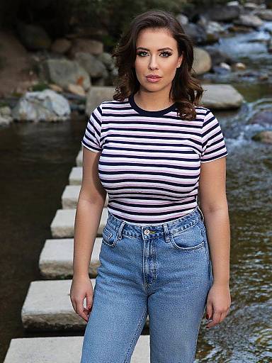 Photograph of a woman with wavy brown hair, wearing a black-and-white striped shirt and high-waisted blue jeans, standing by a rocky