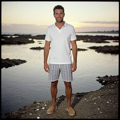 Photograph of a muscular, bearded man in a white polo and striped shorts standing on rocky shoreline at sunset.