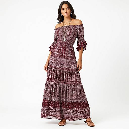 Photograph of a dark-haired woman in an off-shoulder, maroon and white, patterned maxi dress with ruffled sleeves, standing against