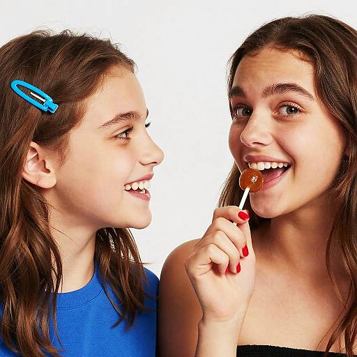Photograph of two smiling young girls with brown hair; one with a blue hair clip, wearing blue shirt, the other licking a lollipop, wearing
