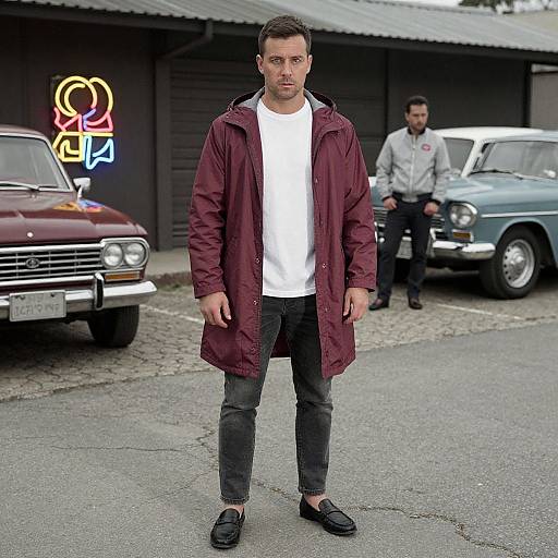 Photograph of a serious-looking man in a white t-shirt and maroon coat, standing in front of vintage cars, with a neon 