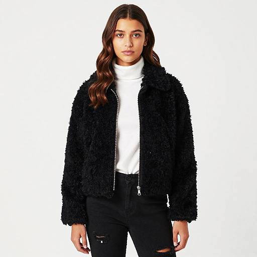 Photograph of a young woman with long brown hair, wearing a black textured jacket, white turtleneck, and black ripped jeans, standing against a