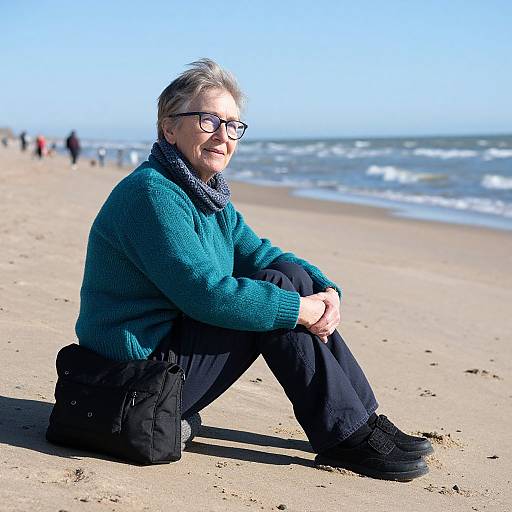 Photograph of an elderly woman with short gray hair, black glasses, teal sweater, black pants, and black boots, sitting on a sunny beach with