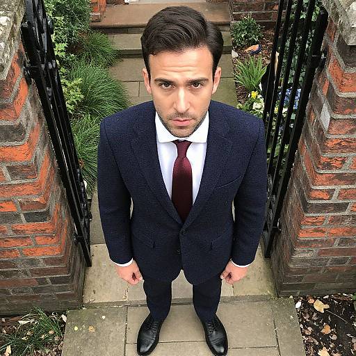 Photograph of a serious-looking man in a black suit, white shirt, and red tie, standing at a brick gate entrance.