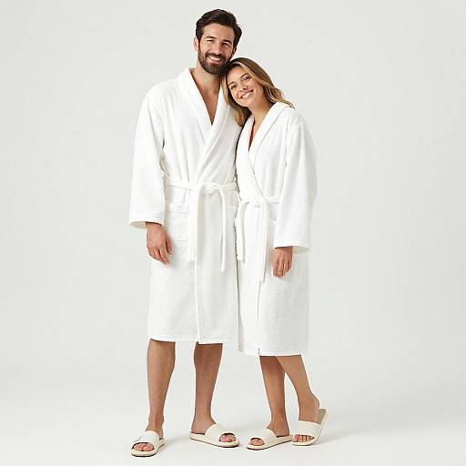 Photograph of a smiling, bearded man and woman standing close together, wearing white bathrobes and white slippers, against a plain white background