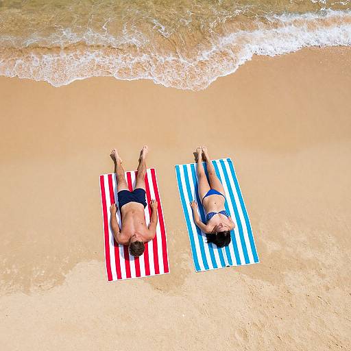 Photograph: Two sunbathers, one on red-striped, the other on blue-striped towel, lying on sandy beach with gentle waves. Both wear