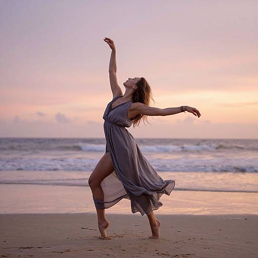Photograph of a woman in a flowing, sleeveless grey dress dancing on a beach at sunset, arms raised gracefully, waves and pink sky in the