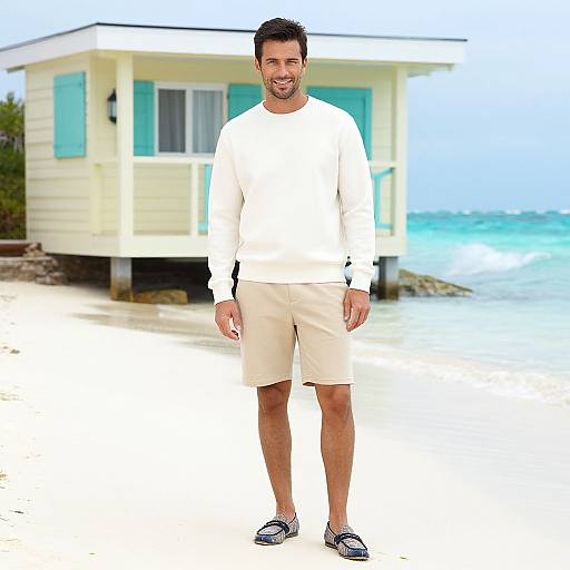 Photograph of a bearded man in a white long-sleeve shirt, beige shorts, and navy slip-ons standing in front of a turquoise-tr