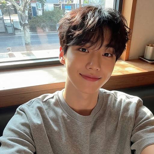 Photograph of a young Asian man with short, dark hair, smiling softly, wearing a gray sweater, seated by a sunlit window.