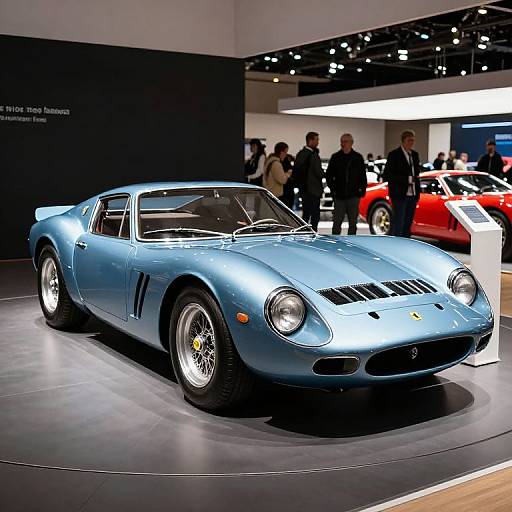 Photograph of a sleek, metallic blue Ferrari 250 GT on display at a car show, with people and a red car in the background.