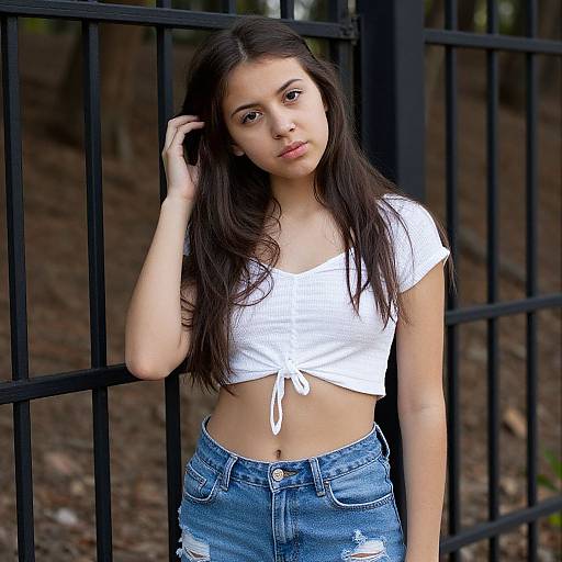Photograph of a young woman with long dark hair, pale skin, and brown eyes, wearing a tied white crop top and distressed blue jeans, standing