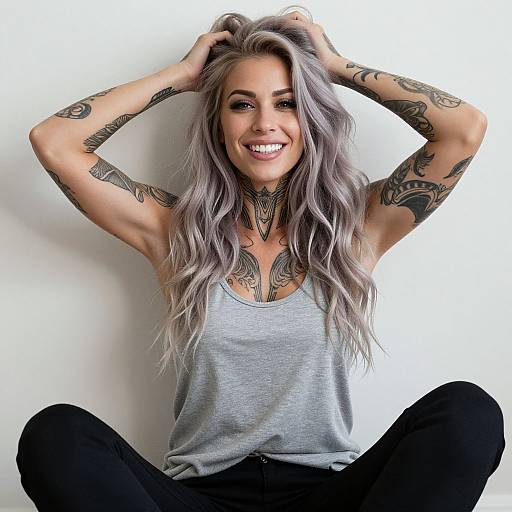 Smiling woman with long, wavy gray hair, tattoos on arms and chest, wearing a gray tank top and black pants, sitting against a white