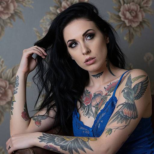 Photograph of a pale-skinned, black-haired woman with extensive colorful tattoos, wearing a blue lace top, against a floral-patterned background.