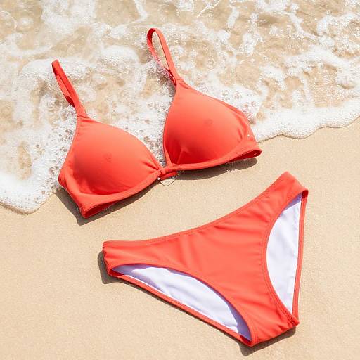 Photograph of a bright red bikini with a triangle top and low-rise bottoms, lying on sandy beach with foamy waves.
