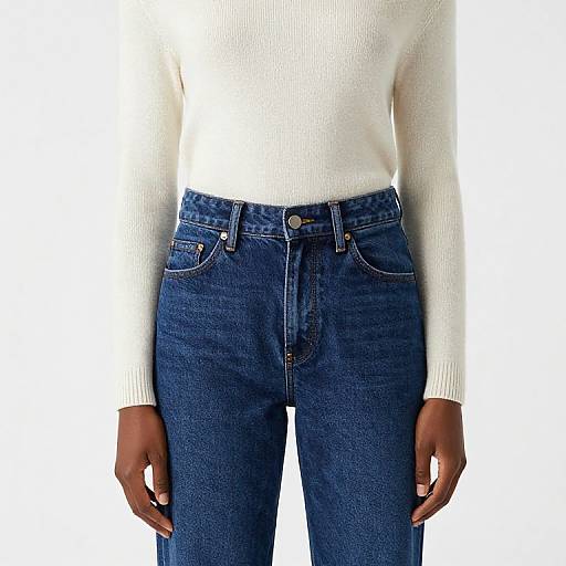 Photograph of a person wearing a white, long-sleeve, ribbed knit sweater and high-waisted, dark blue denim jeans against a
