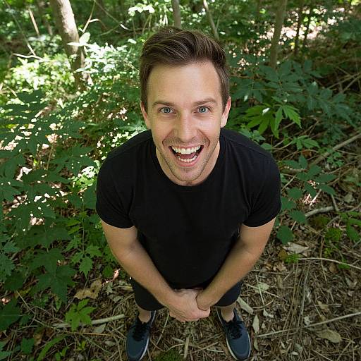 Photograph of a smiling Caucasian man with short brown hair, wearing a black t-shirt and black shoes, standing in a sunlit forest, hands clas