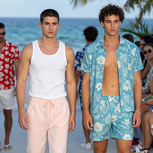 Photograph of two young men on a beach: one in a white tank top and pink shorts, the other in blue floral shorts with an open shirt