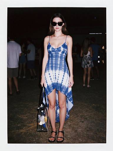 Photograph of a stylish woman in a blue and white tie-dye dress, black sunglasses, holding a handbag, standing on a dark, crowded