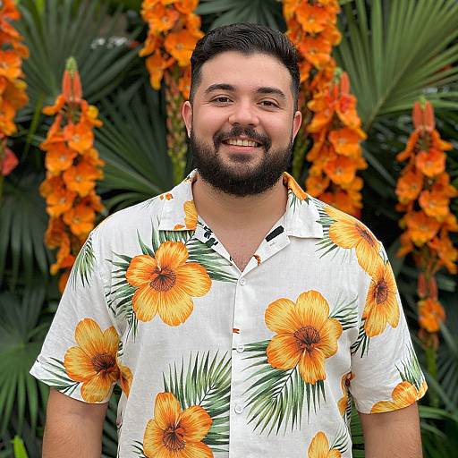 Photograph of a smiling, bearded man with dark hair, wearing a white Hawaiian shirt with large orange flowers, standing in front of vibrant orange and