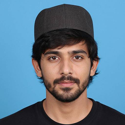 Photograph of a young South Asian man with a black cap, black shirt, and beard, against a bright blue background.