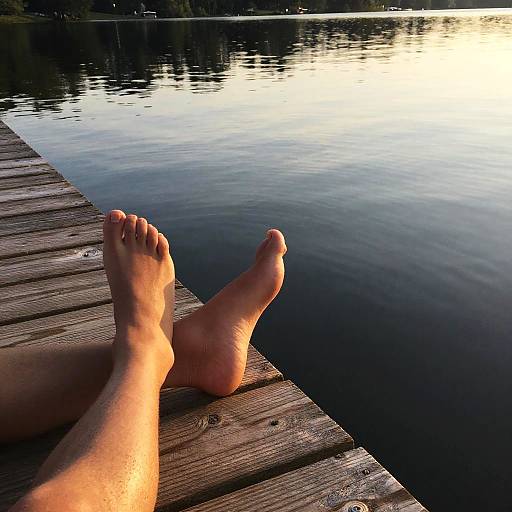 Photograph of two bare feet with light skin tone, resting on a wooden dock, overlooking a calm, reflective lake at sunset.