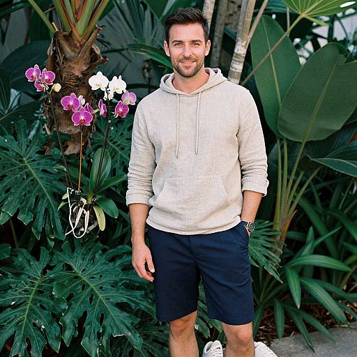 Photograph of a smiling, bearded man in a white hoodie and black shorts, standing in a lush, tropical garden with purple orchids and large