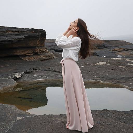 Photograph of a woman with long brown hair, wearing a white blouse and pink flowing skirt, standing on rocky shore with a small pool, hands in