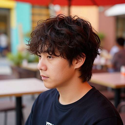 Photograph of an Asian teenage boy with messy black hair, fair skin, wearing a black shirt, in a blurred outdoor café.