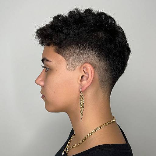 Side profile photograph of a young person with short, curly black hair, undercut, wearing a gold chain and earring, against a plain white background.