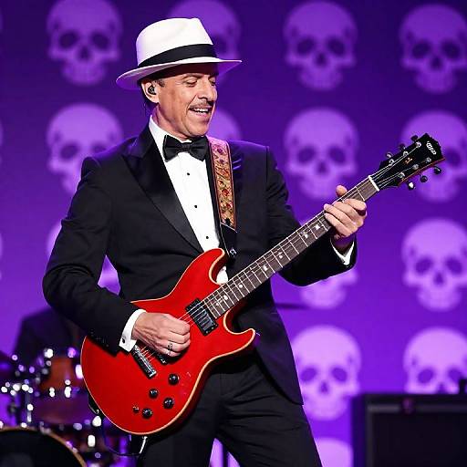 Photograph of a smiling older man in a black tuxedo, white bow tie, and white fedora, playing a red electric guitar on stage