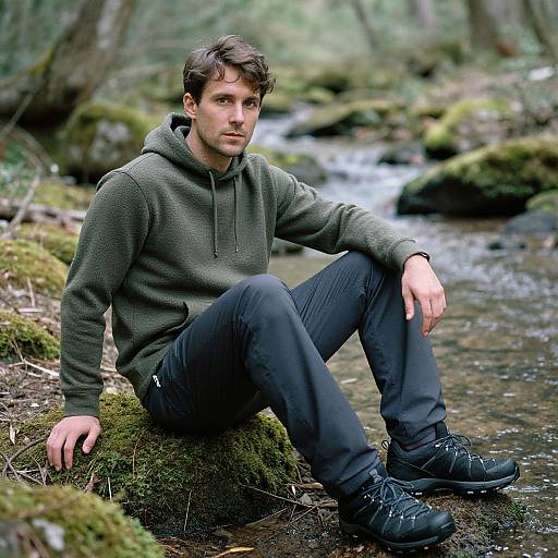 Photograph of a young, handsome man with short brown hair, wearing a green hoodie, black pants, and black shoes, sitting on a moss-covered