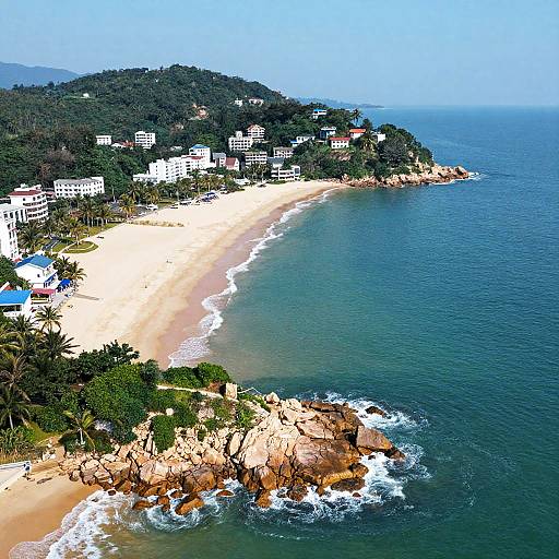 Aerial photograph of a tropical beach with white sand, clear blue ocean, rocky shoreline, lush green hills, and white apartment buildings.