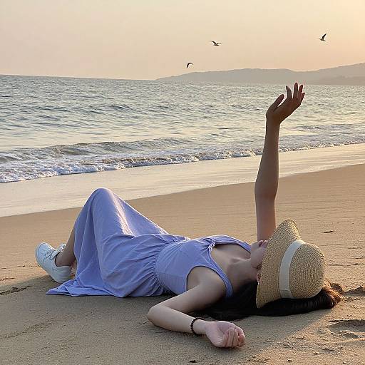 Photograph of a woman with fair skin, wearing a blue dress and straw hat, lying on a beach, arm raised, watching sunset over the ocean