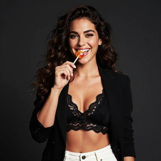 Photograph of a smiling woman with long, wavy brown hair, wearing a black lace bralette, black blazer, and white pants, holding