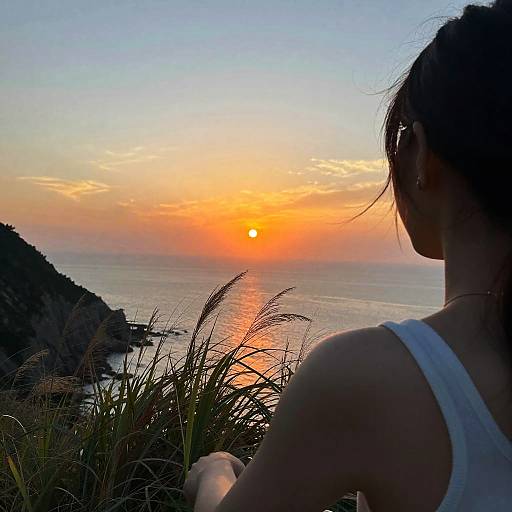 Photograph of a woman with dark hair in a white tank top, silhouetted against a vibrant sunset over a calm ocean and grassy landscape