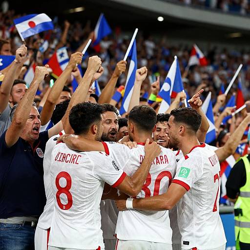 Photograph of four male soccer players in white jerseys with red accents celebrating, surrounded by cheering fans with flags, in a crowded stadium.