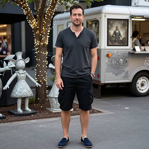 Photograph of a muscular, bearded man in a black polo and shorts, standing on a street, with a food truck and illuminated tree in the