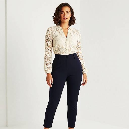 Photograph of a confident woman with curly hair, wearing a white lace blouse and high-waisted black pants, standing against a bright white background.